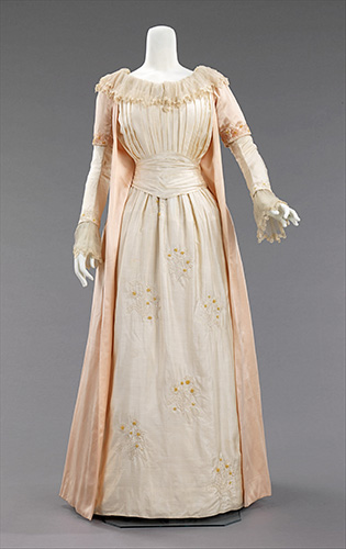 A photograph of an antique silk gown on a mannequin. It is pale peach and cream white, and it has a pleated bodice, a skirt embroidered with daisies, and collar and cuffs fringed with ruffled lace.