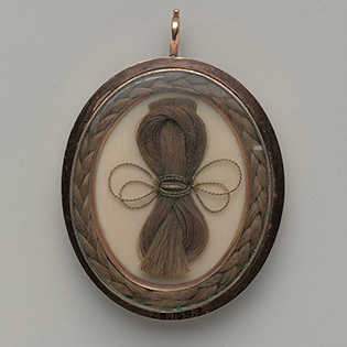 A photograph of a flat, oval pendant containing locks of hair artfully braided, knotted, and tied with a thin string.